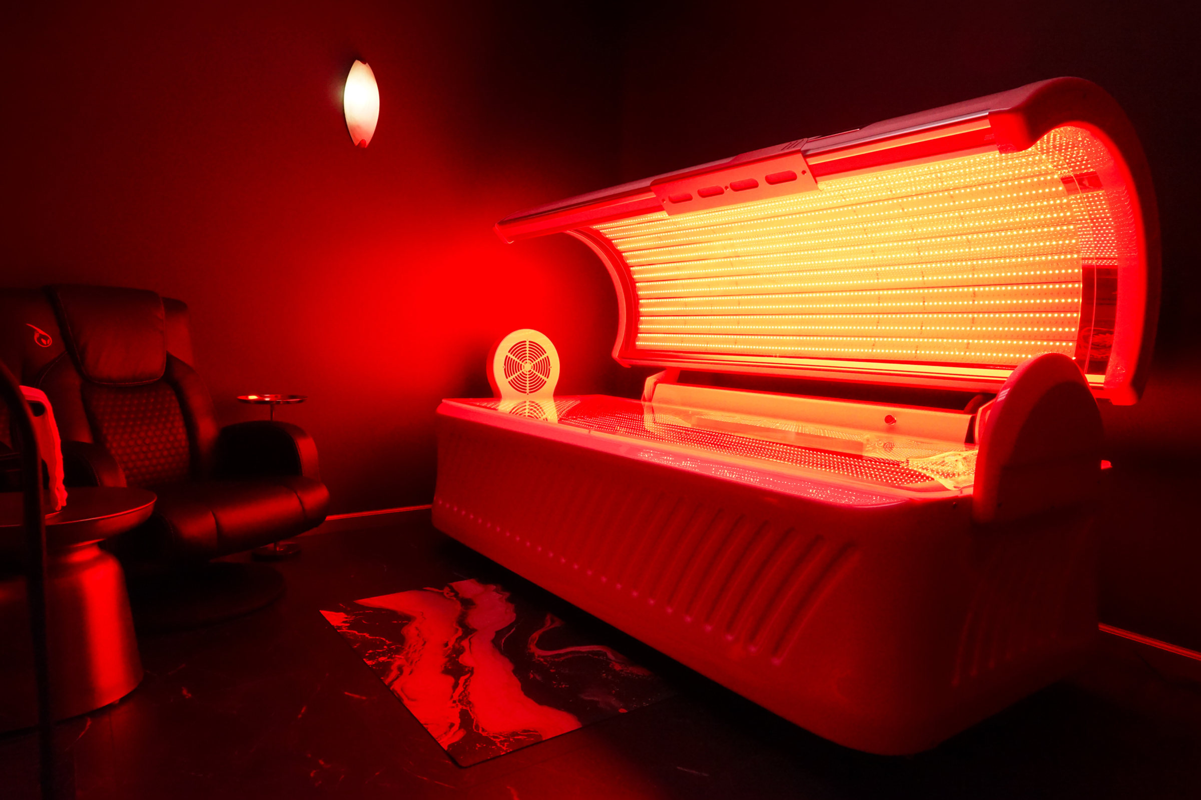 Full-body LED light therapy bed glowing red in a private room with a leather recliner